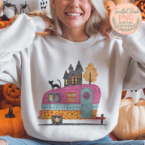Könnte beinhalten: Weißes Sweatshirt mit einem farbenfrohen Wohnwagen-Design, einer schwarzen Katze, einem Spukhaus und einem Herbstbaum. Der Wohnwagen ist rosa, blau und gelb. Das Sweatshirt hat ein Logo mit Text.