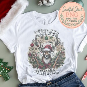 May include: White t-shirt featuring a graphic of a reindeer wearing a Santa hat, with the words "REINDEER HUNTER" above and below the image. The design includes rifles, Christmas ornaments, and a target.