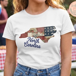 Puede incluir: Camiseta blanca con un diseño de patchwork del contorno del estado de Carolina del Norte. El diseño incorpora patrones rojos, blancos y azules, incluyendo estrellas y estampados florales, con la frase "North Carolina" en azul marino.