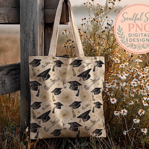 A beige tote bag with a graduation cap pattern is hanging on a wooden fence, with a field of wildflowers and a pink circular logo in the background.