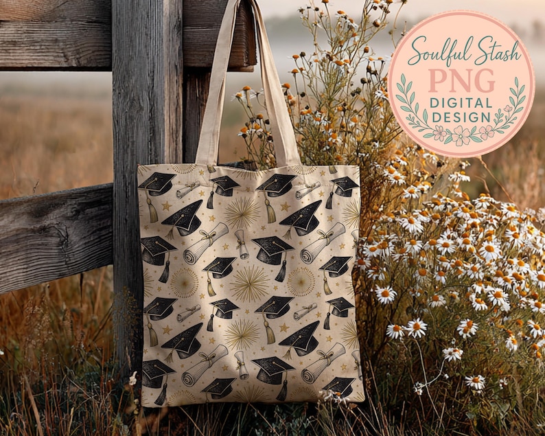 A beige tote bag with a graduation cap pattern is hanging on a wooden fence, with a field of wildflowers and a pink circular logo in the background.
