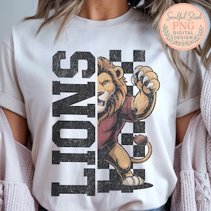 May include: Cream-coloured t-shirt with a distressed graphic of a lion wearing a maroon shirt, and the word "LIONS" in large, black, block letters. A black and white chequered pattern is behind the lion.