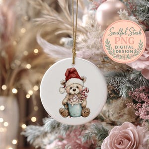 A white ceramic ornament featuring a teddy bear wearing a Santa hat and holding a cup of hot chocolate, hanging from a string against a backdrop of a Christmas tree with pink and white ornaments.