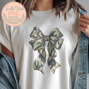 May include: White t-shirt featuring a large bow design in a camouflage pattern of green, brown, and tan. The bow is centered on the shirt. The shirt also has a circular logo with text that reads "Soulful Stash PNG DIGITAL DESIGN".