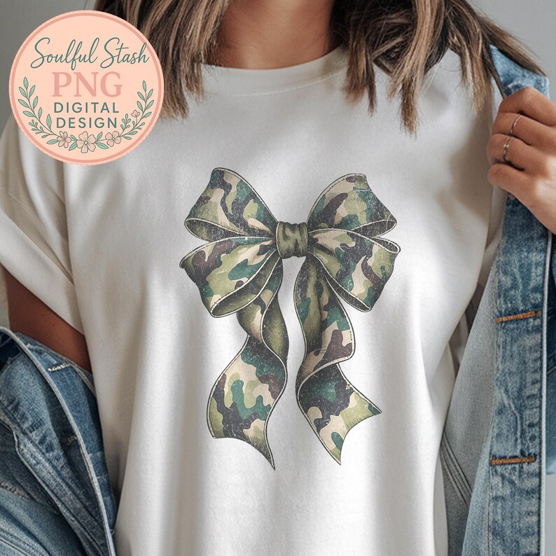 May include: White t-shirt featuring a large bow design in a camouflage pattern of green, brown, and tan. The bow is centered on the shirt. The shirt also has a circular logo with text that reads "Soulful Stash PNG DIGITAL DESIGN".