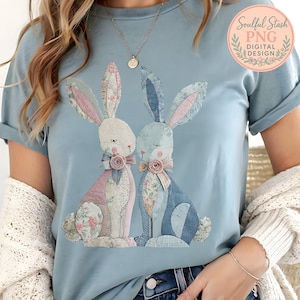 Puede incluir: Camiseta azul claro con un gráfico de dos conejos con cuerpos de patchwork y detalles florales. Los conejos tienen orejas rosas y azules y están adornados con lazos. La camiseta tiene mangas cortas y cuello redondo.