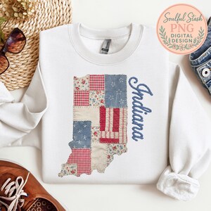 May include: White sweatshirt featuring a patchwork design in the shape of Indiana. The patchwork includes red gingham, floral, and star-patterned fabrics. The word "Indiana" is embroidered in blue script on the right side.