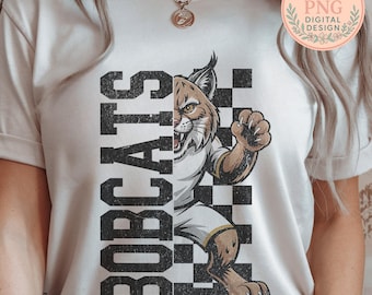 Pode incluir: Camiseta creme com um desenho gráfico de um lince e a palavra "BOBCATS" em fonte vertical e desgastada. O lince está sobre um fundo quadriculado preto e branco, com o logotipo "Soulful Stash PNG DIGITAL DESIGN" no canto superior direito.