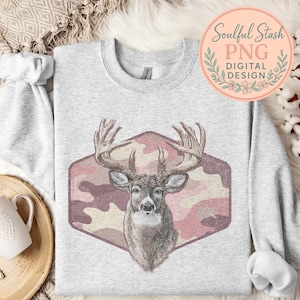 May include: A light gray sweatshirt featuring a deer head graphic within a pink camouflage hexagon. The deer has large antlers and a detailed face. The sweatshirt also has the text "Soulful Stash PNG DIGITAL DESIGN" in a circular design.