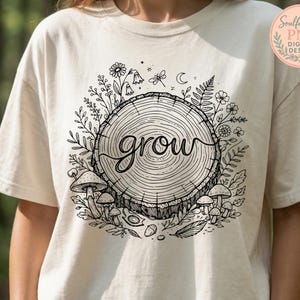 May include: Cream-colored t-shirt featuring a black and white illustration of a tree stump with the word "grow" in cursive. The design includes mushrooms, flowers, and foliage around the stump. The shirt has short sleeves.