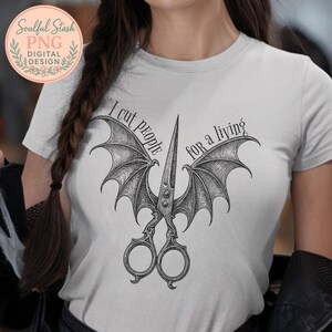 May include: A pale grey t-shirt with a black graphic of scissors with bat wings. The text "I cut people for a living" is arched around the scissors. The shirt has a round neckline and short sleeves.