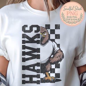 May include: White t-shirt featuring a distressed graphic of a hawk wearing a white shirt, set against a black and white checkered background. The word "HAWKS" is printed vertically in bold, black letters. The design includes the text "Soulful Stash PNG DIGITAL DESIGN".