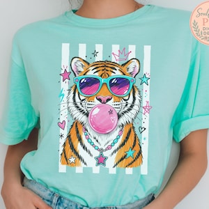A person wearing a turquoise t-shirt with a graphic design of a tiger blowing a bubble gum bubble, surrounded by colorful stars and hearts.