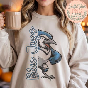 May include: Cream-colored sweatshirt featuring a blue jay mascot graphic. The graphic includes the text "Blue Jays" in a stylized font. The blue jay is depicted in shades of blue, white, and gray, with a distressed, vintage look.