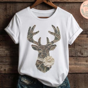 May include: White t-shirt featuring a deer head design. The deer head is made of camouflage fabric with a cream-colored flower and ribbon. The shirt is hanging on a wooden hanger against a wooden background. The text "Soulful Stash PNG Digital Design" is in the upper right corner.