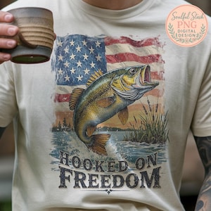 May include: Cream-colored t-shirt featuring a graphic of a largemouth bass leaping from water, set against a distressed American flag. The text "HOOKED ON FREEDOM" is below the fish. A floral design with the text "Soulful Stash PNG Digital Design" is in the upper right.