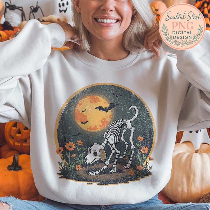 May include: Cream-colored sweatshirt featuring a Halloween-themed graphic. The design includes a dog skeleton, a full moon, bats, and flowers. The sweatshirt has a round neck and long sleeves. The text "Soulful Stash PNG Digital Design" is in the upper right corner.