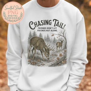May include: A white crewneck sweatshirt featuring a graphic print of two deer in a forest setting. The text reads "CHASING TAIL! FRIENDS DON'T LET FRIENDS RUT ALONE." The design includes detailed illustrations of trees, fallen leaves, and a buck with antlers.