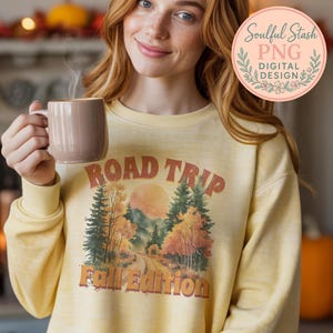 May include: A light yellow sweatshirt with a "Road Trip Fall Edition" graphic. The graphic features a road winding through a forest with fall foliage and a setting sun. A brown mug is held in the hand.
