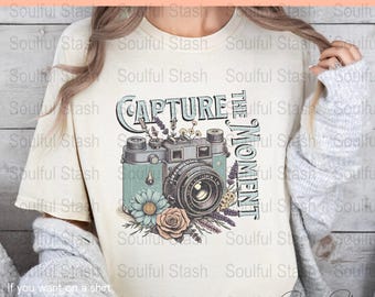 May include: A beige t-shirt with a vintage camera graphic and the text "Capture the Moment". The camera is surrounded by flowers and greenery.