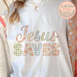 May include: White sweatshirt with the words "Jesus Saves" in a floral and gingham pattern. The word "Jesus" is in a script font with a floral pattern, and "Saves" is in a block font with a gingham pattern.