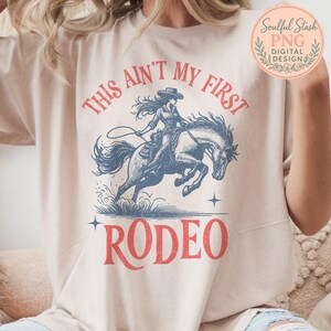 以下が含まれることがあります： ベージュ色のTシャツ。カウガールが馬に乗るヴィンテージスタイルのイラストが描かれています。「THIS AIN'T MY FIRST RODEO」の文字が赤色でグラフィックの上と下にプリントされています。デザインは使い古されたような外観です。