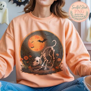 May include: Peach-colored sweatshirt featuring a Halloween-themed graphic. The graphic depicts a dog skeleton in a field of flowers, with a full orange moon and bats in the background. The sweatshirt has long sleeves and a crew neck.