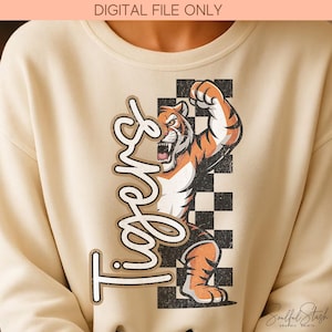May include: Cream-colored sweatshirt featuring a cartoon tiger illustration with orange and black stripes, flexing its arm. The word "Tigers" is written in a stylized font. A black and white checkered pattern is behind the tiger.