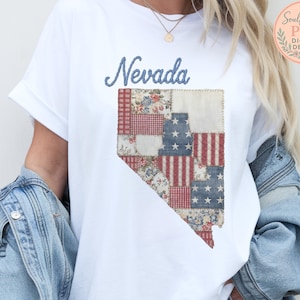 May include: White t-shirt with a patchwork design of the Nevada state outline. The design incorporates red, white, and blue patterns, including stars and stripes. The word "Nevada" is written above in blue script.