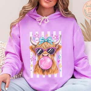 A woman wearing a purple hoodie with a graphic of a horned animal, likely a yak or a cow, wearing sunglasses and blowing a bubble with a pink bubblegum bubble.