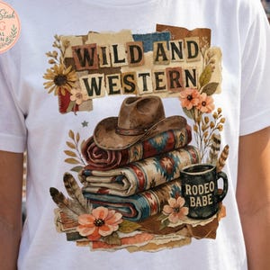May include: White t-shirt featuring a "Wild and Western" design. The graphic includes a cowboy hat, stacked blankets with a Southwestern pattern, a coffee mug that says "Rodeo Babe", and floral accents. The design is a digital print.