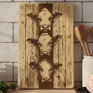 May include: A rectangular wooden cutting board with a vertical design of three cow faces. The cow faces are etched into the wood, with a light brown background and darker brown details. The board is displayed in a kitchen setting.