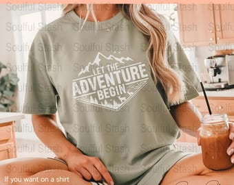May include: A green t-shirt with a white graphic design that says "Let the Adventure Begin" with a mountain range, trees, and a deer. The design is in a diamond shape.