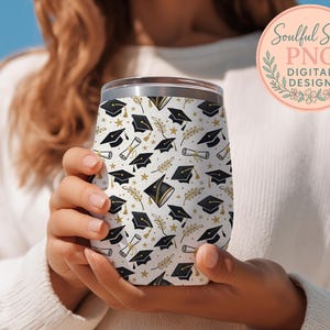A person is holding a white wine glass with a pattern of black graduation caps and laurels on it.