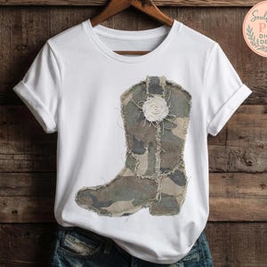 May include: White t-shirt featuring a camouflage cowboy boot design with a white floral accent. The boot design incorporates shades of green, brown, and tan. The shirt is displayed on a wooden background.