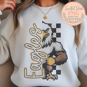 May include: White sweatshirt featuring a distressed graphic of a fierce eagle with the word "Eagles" in a yellow and black font. The eagle is set against a black and white checkered background. The design includes the text "Soulful Stash PNG Digital Design".