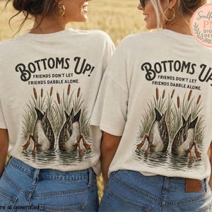 May include: An off-white t-shirt featuring a graphic on the back with two ducks diving into water among cattails. The text reads "BOTTOMS UP! FRIENDS DON'T LET FRIENDS DABBLE ALONE." The design is rendered in black, brown, and green tones.