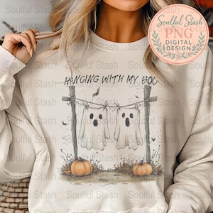 May include: Cream-colored sweatshirt with a Halloween-themed graphic. The design features two ghost figures hanging on a clothesline with the text "Hanging with my Boo". Two orange pumpkins are at the bottom.