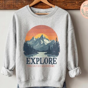 May include: A heather gray sweatshirt with a mountain and sunset graphic. The word "EXPLORE" is printed across the bottom, with the phrase "EVERY DAY IS A NEW ADVENTURE" below it. The sweatshirt is hanging on a wooden hanger.
