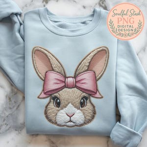 Op de afbeelding: Een lichtblauw sweatshirt met een geborduurd konijnengezicht en een roze strik. Het konijn heeft bruine vacht, roze oren en grote bruine ogen. De sweatshirt heeft opgerolde mouwen en de tekst "Soulful Stash PNG DIGITAL DESIGN" in een cirkelvormig logo.