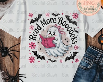 Pode incluir: Camiseta branca com um fantasma de desenho animado lendo um livro rosa, rodeado por morcegos, flores e o texto "Read More Boooooks". O design é em preto e rosa, perfeito para o Halloween ou amantes de livros.