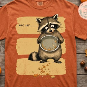 Könnte beinhalten: Ein orangefarbenes T-Shirt mit einer Grafik eines Waschbären, der eine leere Kuchenform hält, Krümel im Gesicht hat und den Text Well, shit....... Der Waschbär ist über drei horizontalen beigefarbenen Pinselstrichen abgebildet.