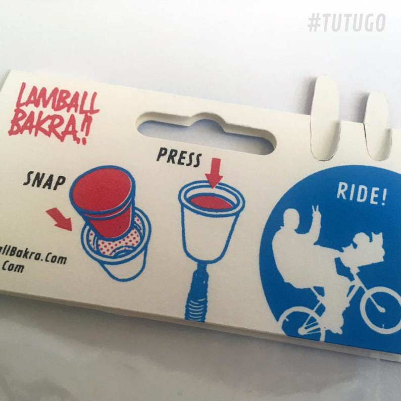 May include: A white cardboard package with instructions for a toy. The package has a red and white cup with the words "SNAP" and "LAMBALL BAKRA!" The package also has a blue circle with a white silhouette of a person riding a bicycle with the words "RIDE!" and "PRESS".