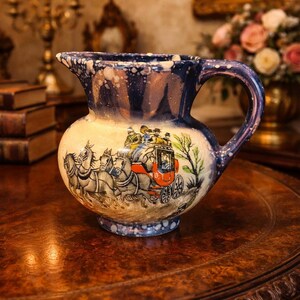 May include: A ceramic pitcher featuring a blue and white speckled glaze on the rim and handle. The body displays a detailed illustration of a horse-drawn stagecoach with passengers, rendered in shades of brown, red, and green on a cream background.