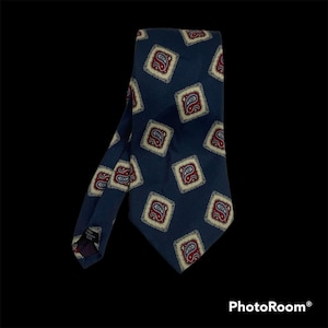 Può includere: Una cravatta blu navy caratterizzata da un motivo ripetuto di cornici quadrate. Ogni quadrato contiene un disegno paisley rosso e bianco su sfondo color crema. La cravatta presenta un'estremità a punta e un'etichetta che indica 100% seta.