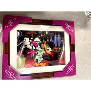 Könnte beinhalten: Ein gerahmtes Animations-Cel mit Woody Woodpecker in einem Laborkittel und rosa Handschuhen, der eine grüne Flüssigkeit aus einem Reagenzglas in ein Becherglas gießt. Der Hintergrund zeigt ein Labor mit Regalen voller Glaswaren und Laborgeräten.