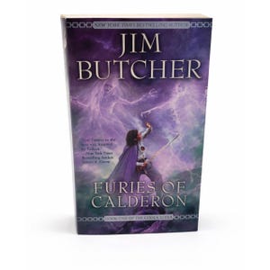 Puede incluir: Un libro de tapa dura titulado "Furies of Calderon" de Jim Butcher. La portada presenta una ilustración de fantasía con una figura sosteniendo una espada, sobre un fondo púrpura y azul con figuras fantasmales. El libro es parte de la serie Codex Alera.