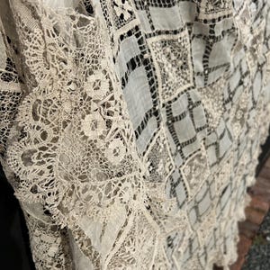 Peut inclure: Un textile blanc cassé présentant un patchwork complexe de panneaux de dentelle complexes et de sections de tissu carrées. Le design intègre des motifs de broderie variés, incluant des fleurs et des grilles géométriques, créant une surface texturée.
