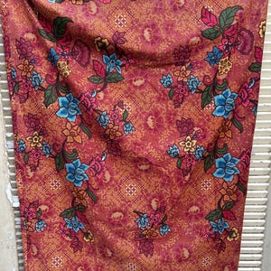 May include: A rectangular fabric textile featuring a dense, intricate floral pattern in shades of deep red, orange, and gold. The design includes clusters of blue, yellow, and pink flowers with green leaves, set against a detailed, repeating geometric background.