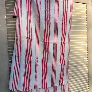 May include: A rectangular textile piece featuring a pattern of vertical red stripes on an off-white background. The fabric displays visible signs of wear, including small frayed edges, loose threads, and faint yellowish discolouration spots.